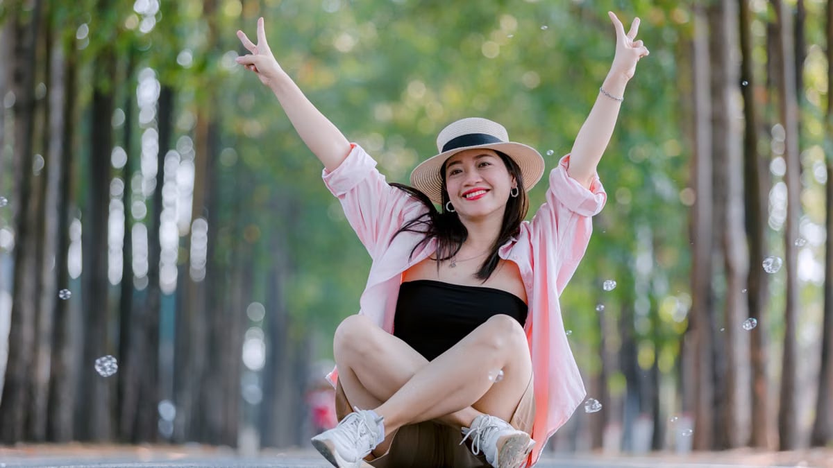 Smiling woman sitting cross-legged on a scenic tree-lined road, wearing a hat and raising peace signs.